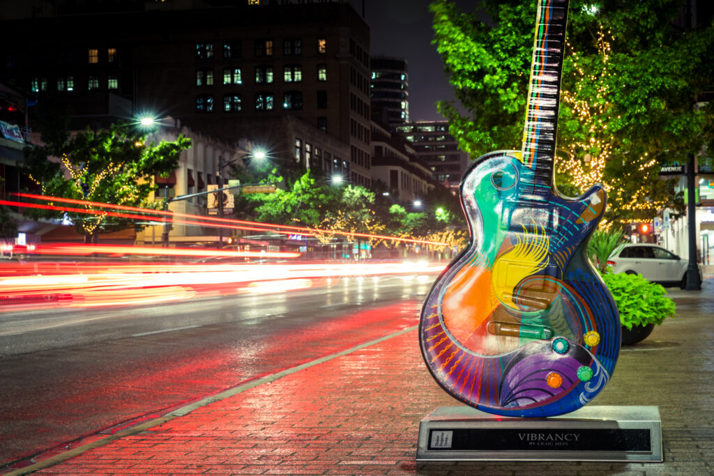 A dark street in downtown Austin Texas, is lit up by car lights speeding by a colorfully-painted guitar, which highlights the city's love for music.